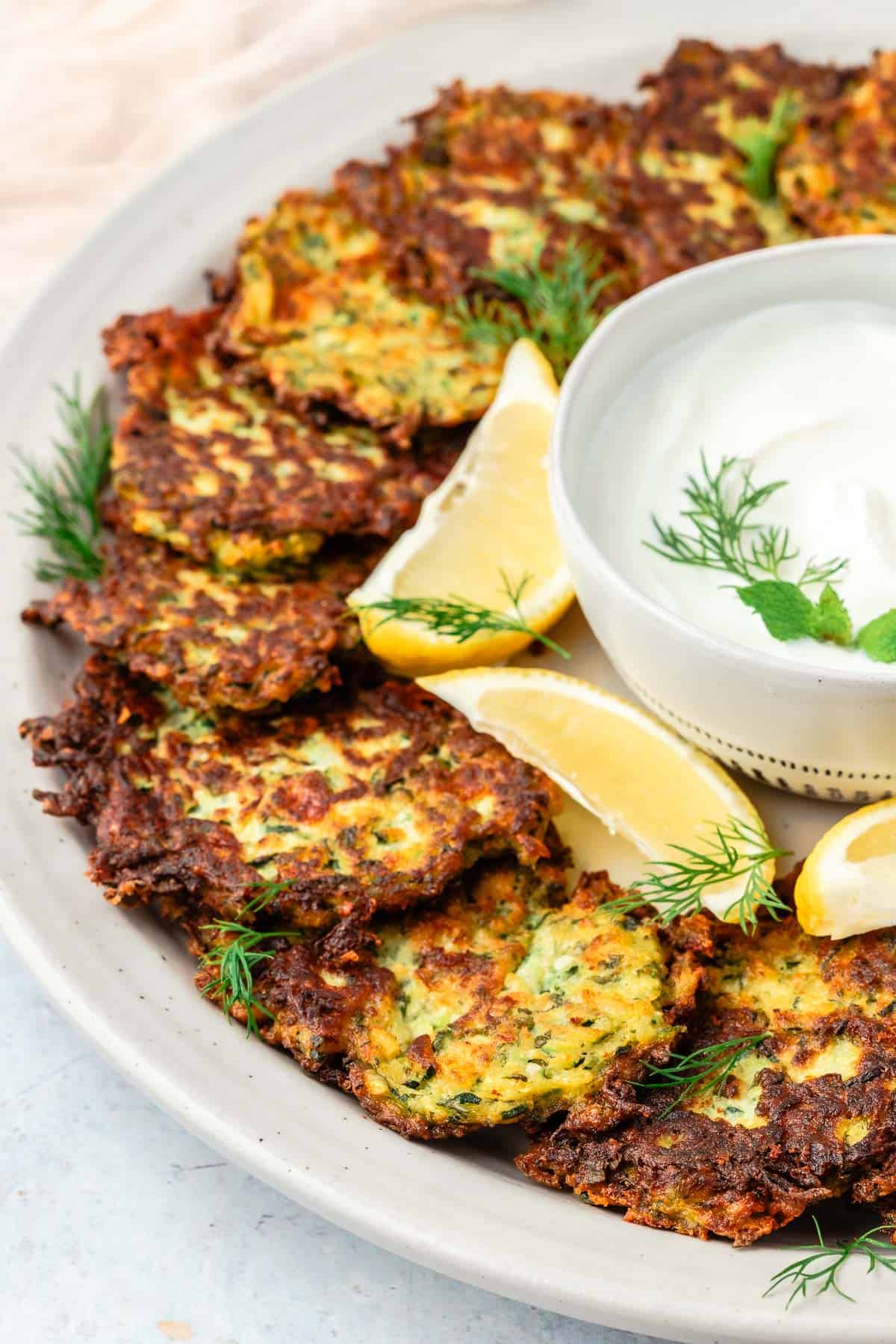 Zucchini fritters on a large platter with a small bowl of greek yogurt and lemon wedges in the center.