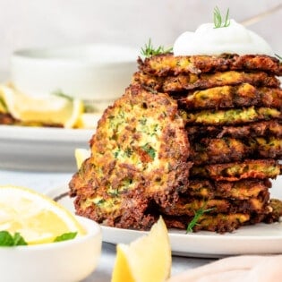 A stack of zucchini fritters with a dollop of Greek yogurt on top and lemon wedges on the side.