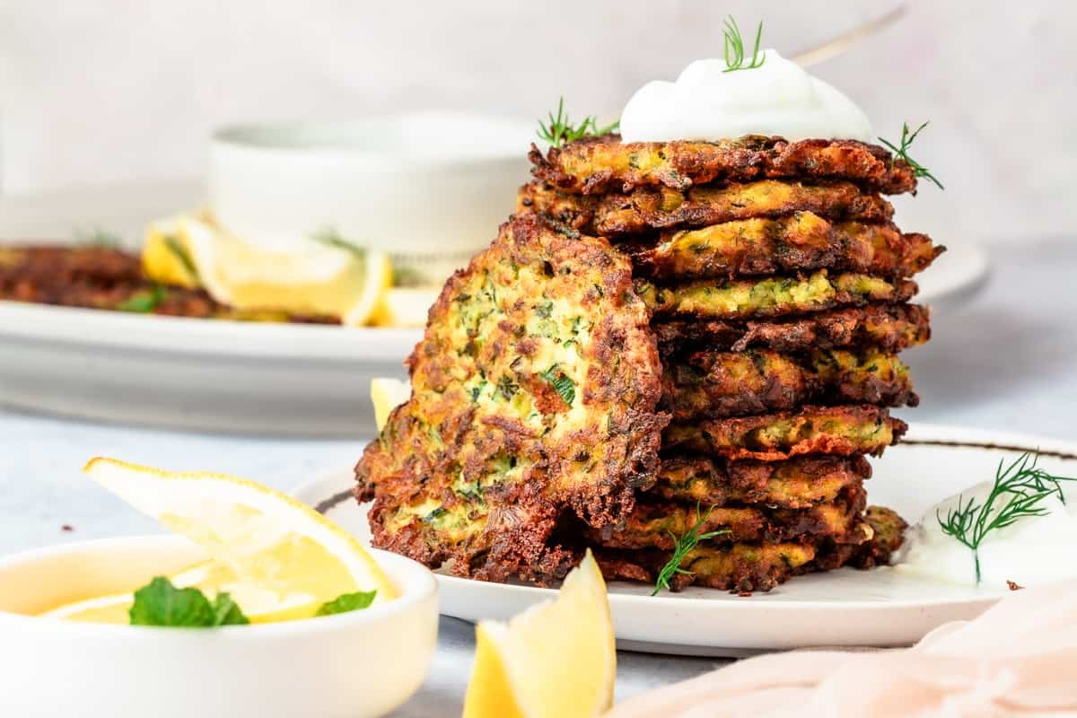 A stack of zucchini fritters with a dollop of Greek yogurt on top and lemon wedges on the side.