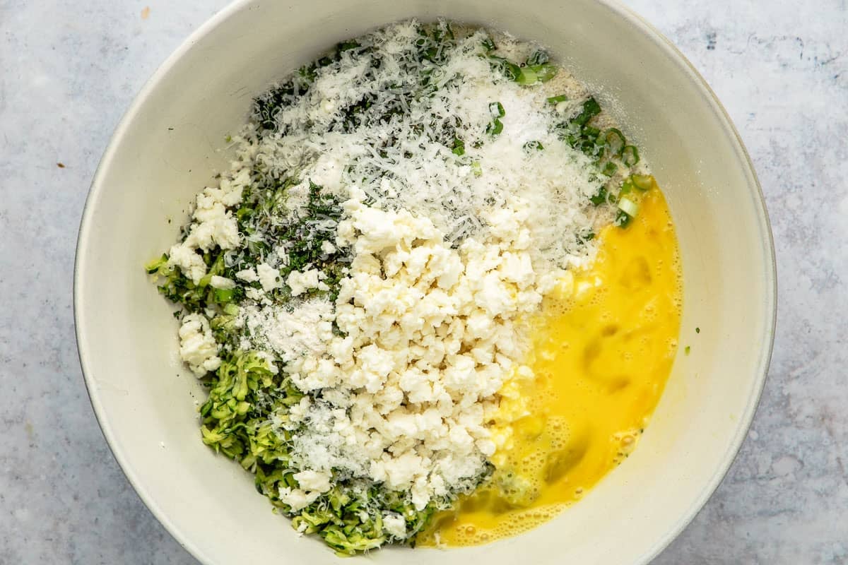 Unmixed zucchini batter in a bowl, including egg, feta cheese, green onion, and shredded zucchini.