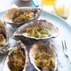 close up of baked oysters with crispy lemon and parsley breadcrumbs on a serving plate with salt, lemon wedges and forks, next to various table settings.