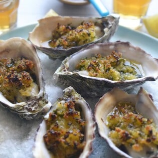 baked oysters with crispy lemon and parsley breadcrumbs on a serving platter with various table setting in the background.