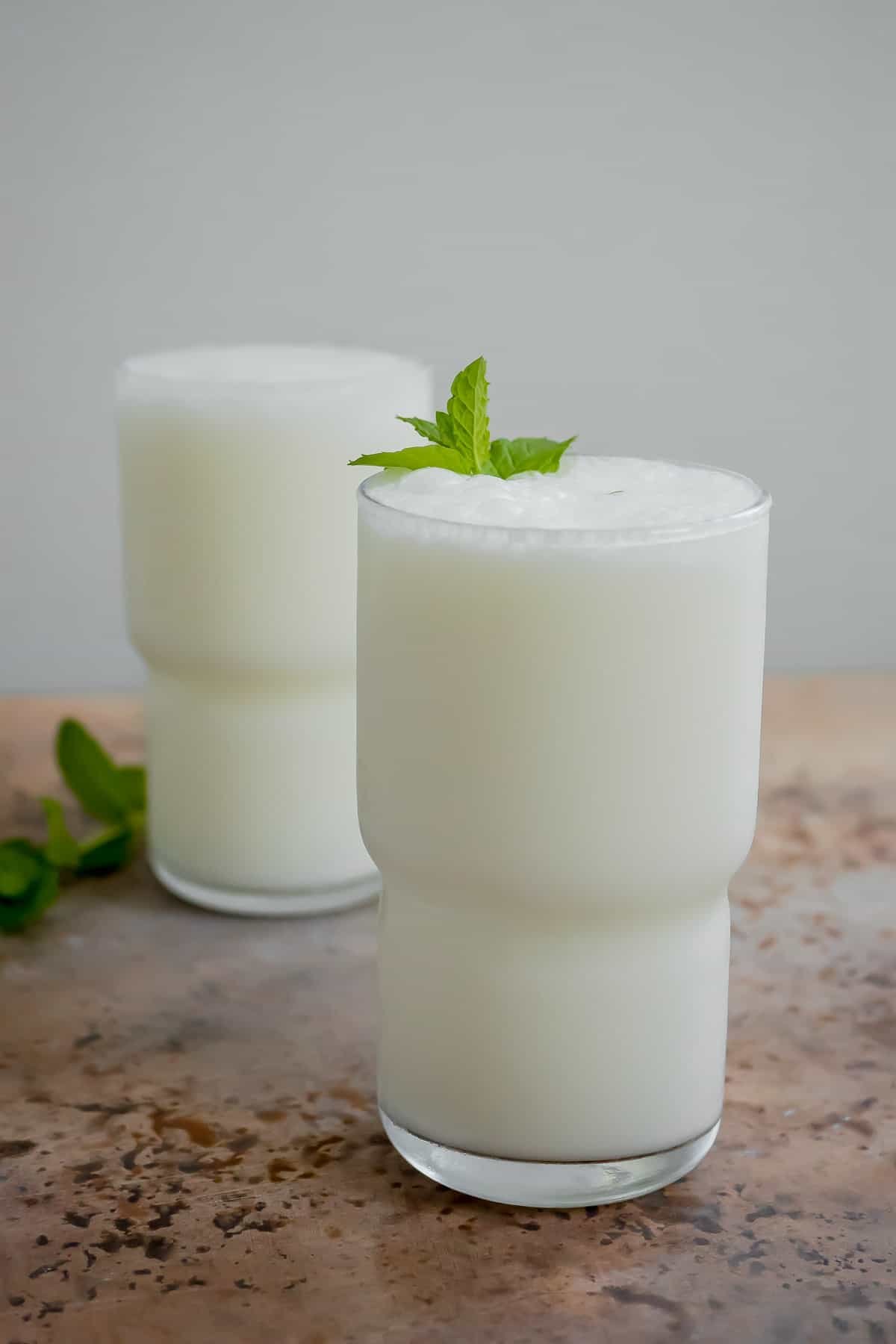 an ayran turkish yogurt drink in a glass garnished with mint leaves, with another glass of ayran in the background.