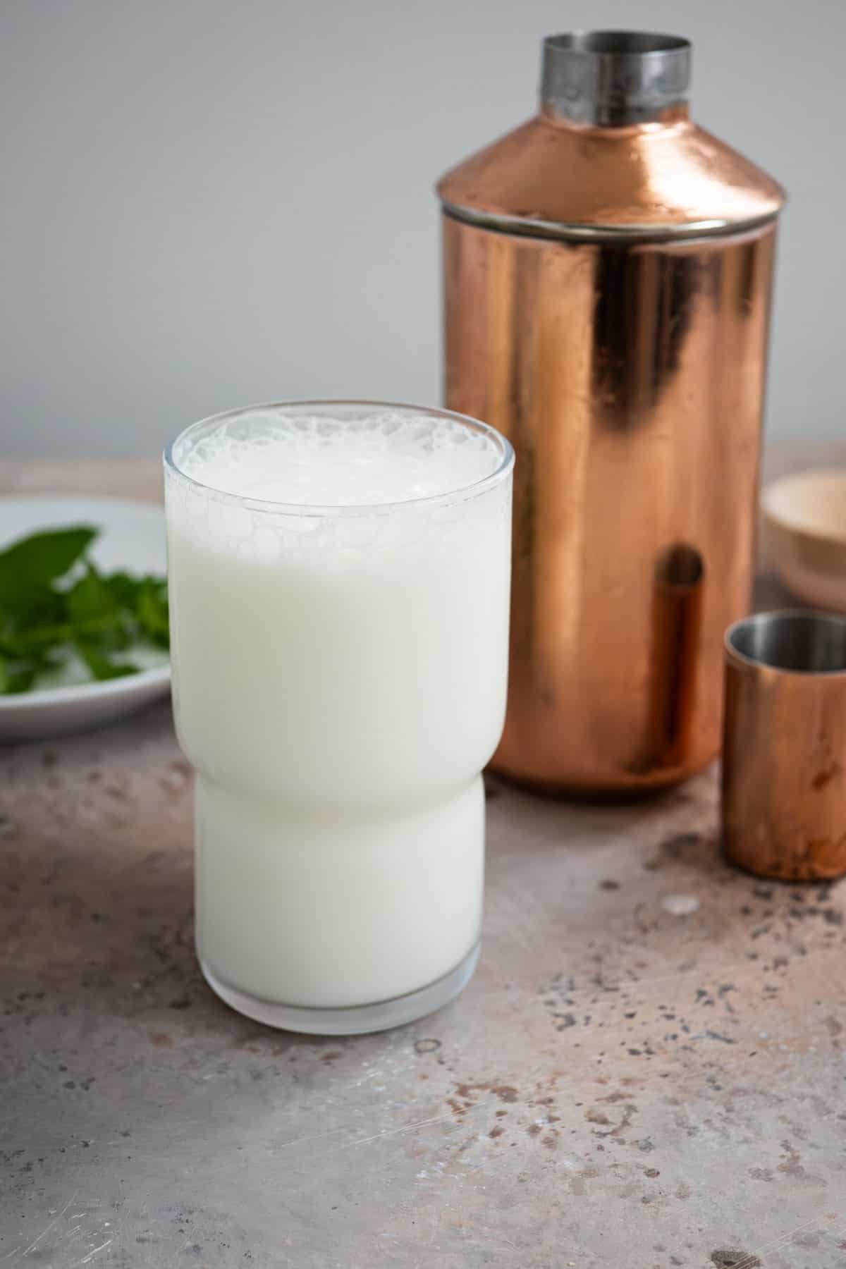 an ayran turkish yogurt drink in a glass in front of a cocktail shaker and a plate of mint leaves.