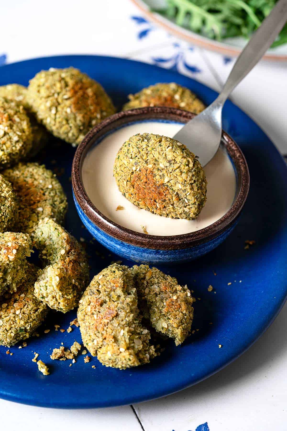 a baked falafel being dipped into a small bowl of tahini sauce on a plate filled with baked falafel.