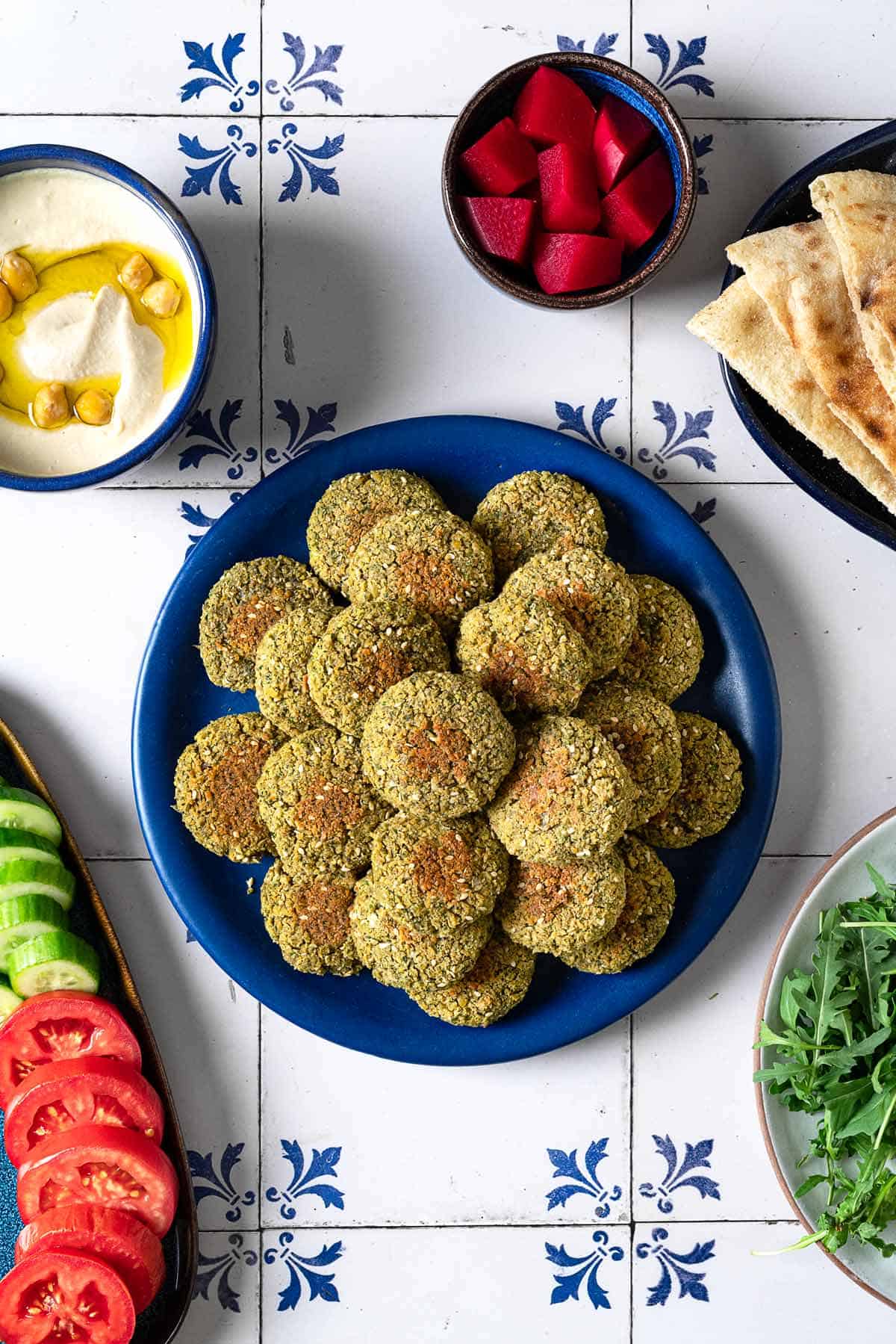 overhead photo of a blue plate full of baked falafel surrounded by a plate of cucumbers and tomatoes, a bowl of hummus, a bowl of pickled turnips, a plate of pita and a bowl of arugula.