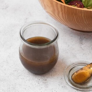 The balsamic vinaigrette recipe in a jar next to a small serving spoon and a bowl of salad.