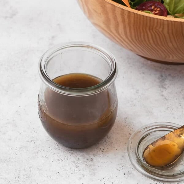 The balsamic vinaigrette recipe in a jar next to a small serving spoon and a bowl of salad.