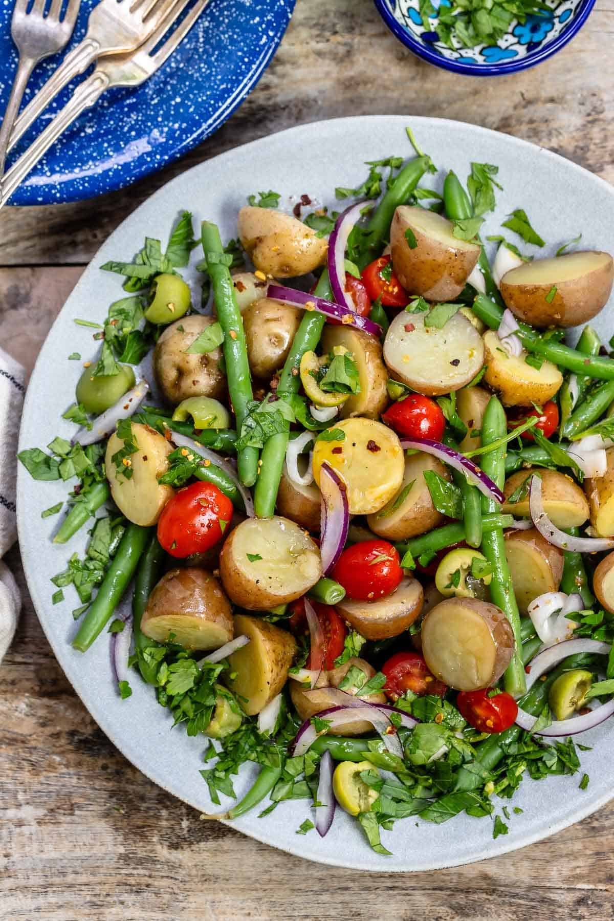 overhead photo of Italian potato salad on a serving platter next to a stack of blue plates with 3 forks, and a small bowl of chopped parsley.