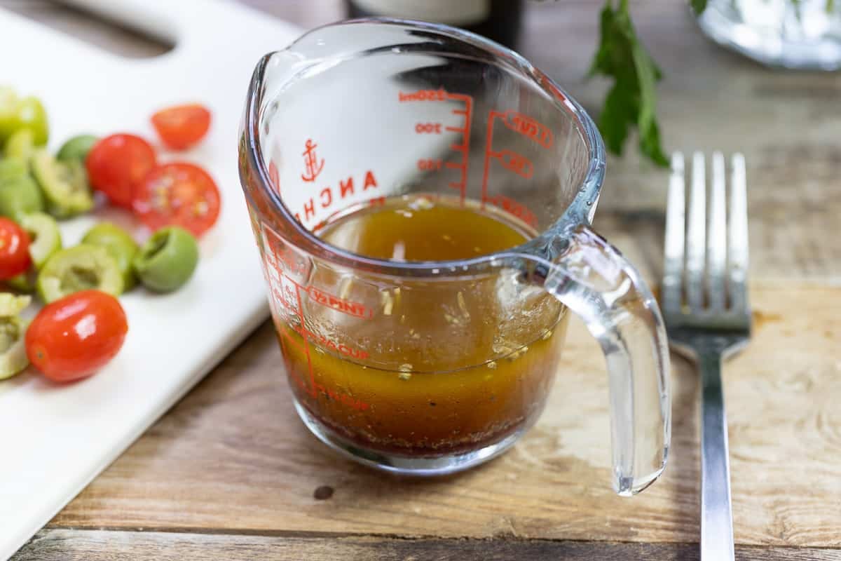 a close up of a measuring cup with salad dressing next to a fork and a cutting board with sliced cherry tomatoes and green olives.
