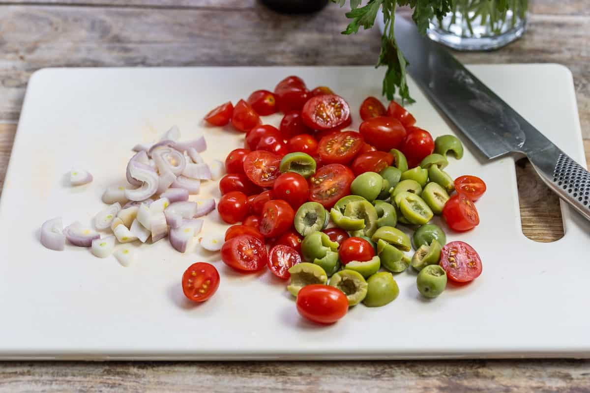 sliced shallots, cherry tomatoes and green olives on a cutting board with a knife.