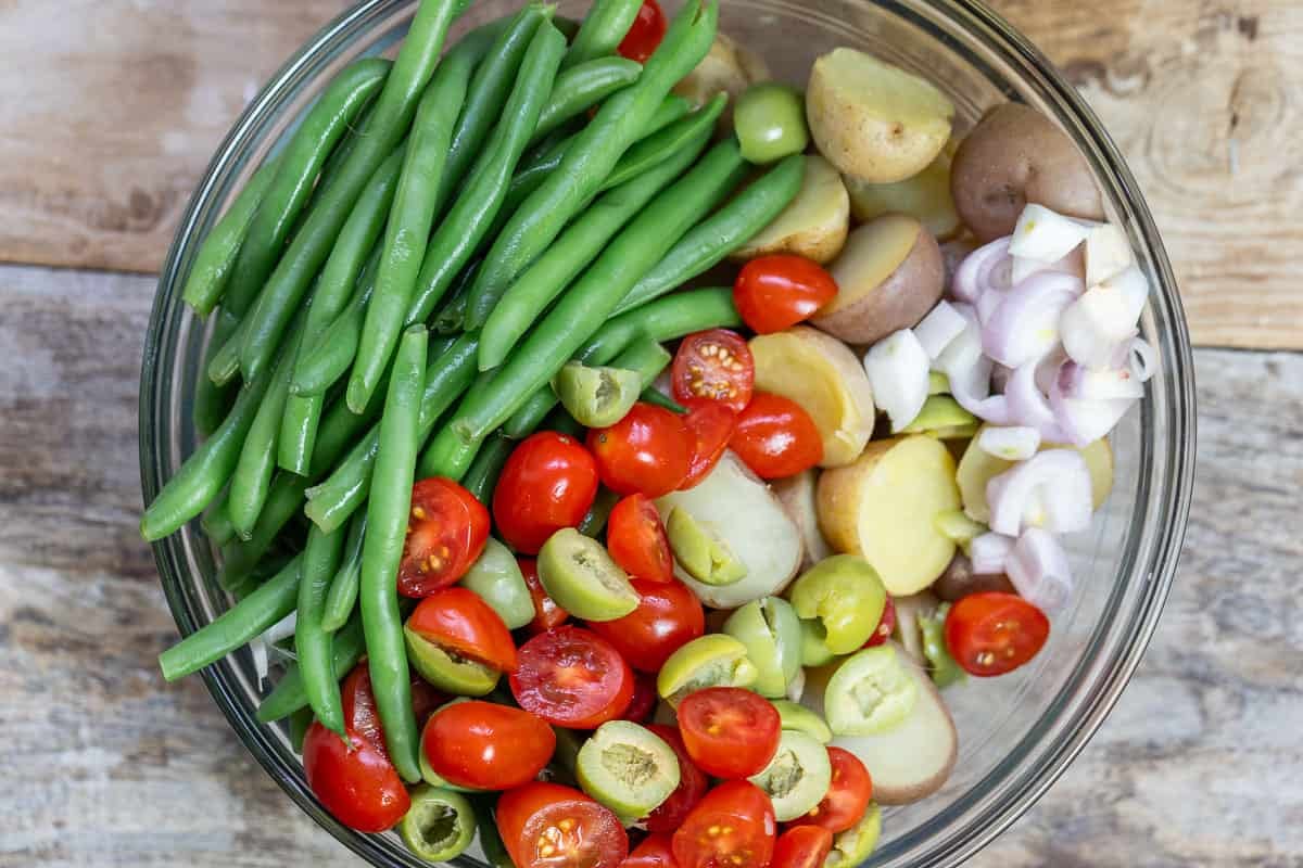 overhead photo of sliced red shallots, green olives and tomatoes in a glass mixing bowl with green beans and halved baby potatoes.