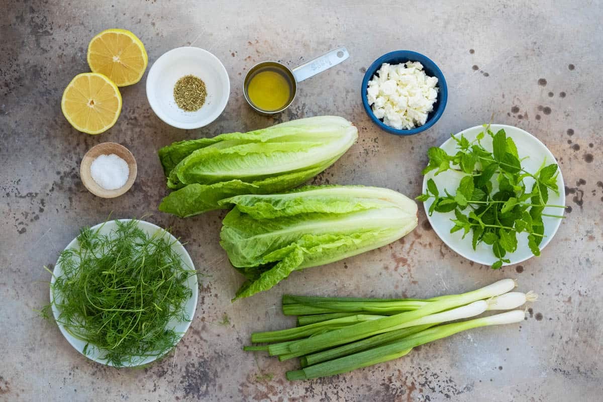 ingredients for maroulosalata including romaine lettuce, green onions, dill, mint, feta cheese, lemon, salt, oregano, and olive oil.