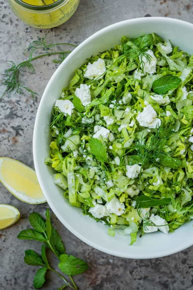 overhead photo of maroulosalata in a white serving bowl, next to a jar of dressing, sliced lemons, dill and mint.