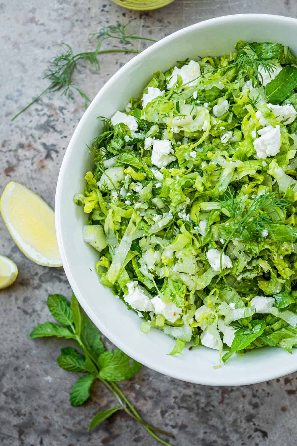 overhead photo of maroulosalata in a white serving bowl, next sliced lemons, dill and mint.