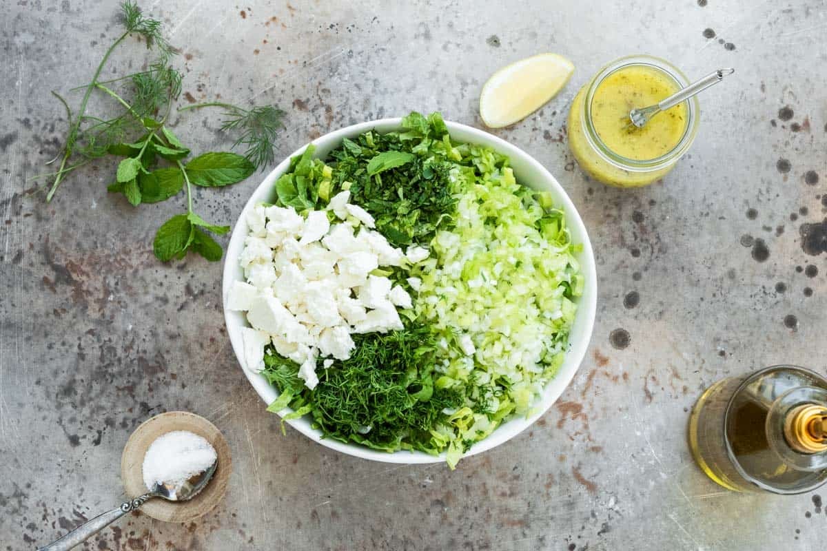 a serving bowl with chopped romaine, green onions, crumbled feta, mint, and dill next to a jar of salad dressing with a whisk, a lemon wedge, sprigs of mint and dill, a bowl of salt with a spoon and a small jug of olive oil.