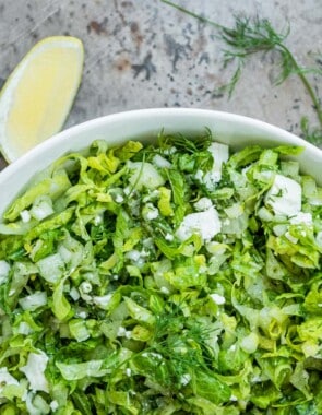 maroulosalata in a white serving bowl next to sprigs of mint and dill, lemon wedges and a bowl of salt with a spoon.