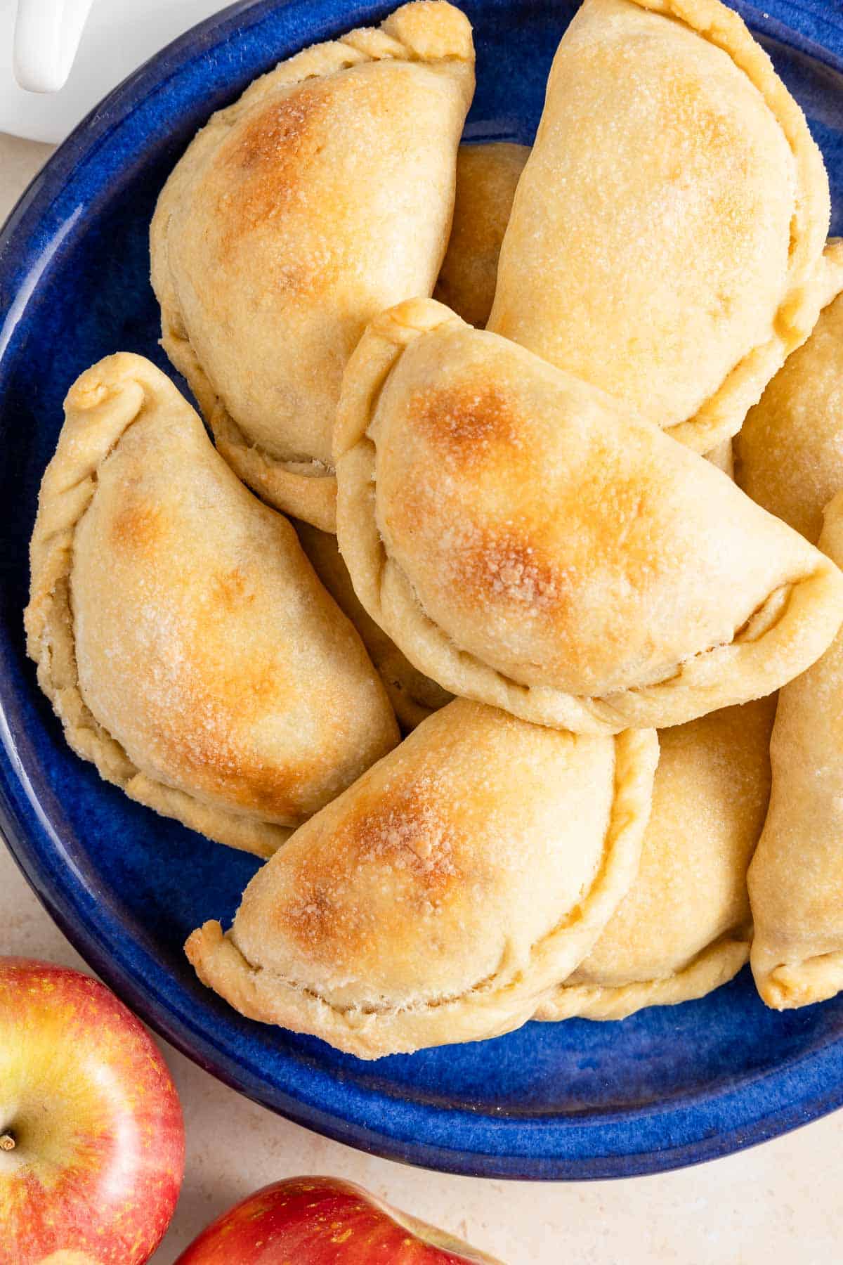 a stack of baked apple empanadas on a blue serving plate next to two apples.