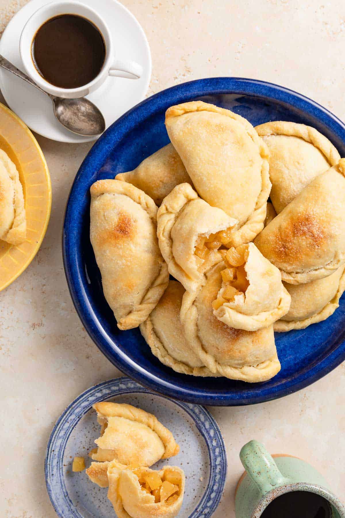 a baked apple empanada cut in half on a stack of other apple empanadas on a blue serving plate, next to a cup of coffee and a plate with one apple empanada on it, also cut in half.