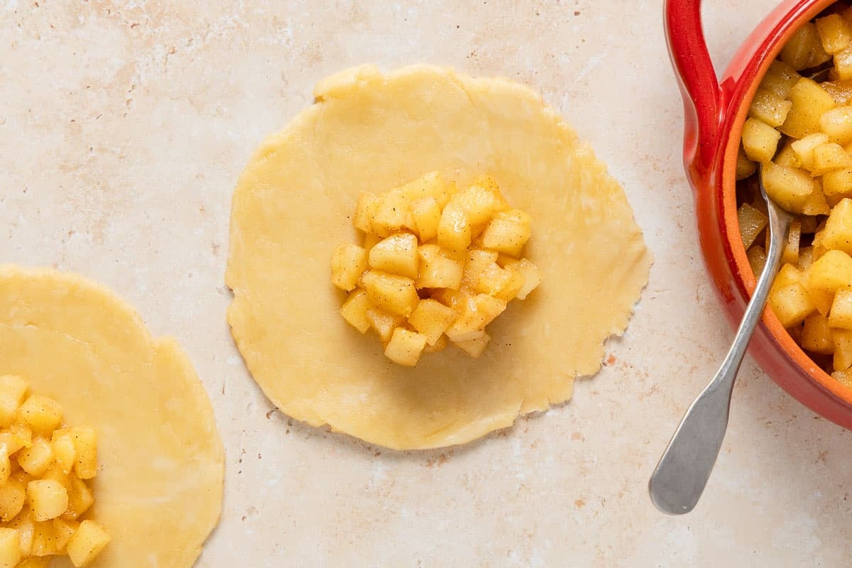 apple filling on two flat rounds of apple empanada dough next to a bowl of apple filling with a spoon.