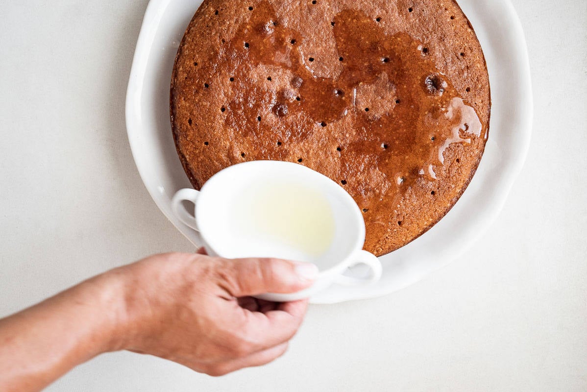 Unglazed Persian love cake with holes poked into the top and a person pouring lemon drizzle over top.