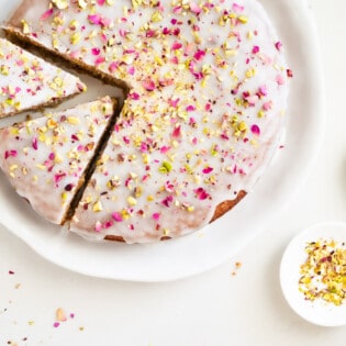 Overhead shot of Persian love cake on a platter with two slices cut out and rose petals and pistachios on the side.