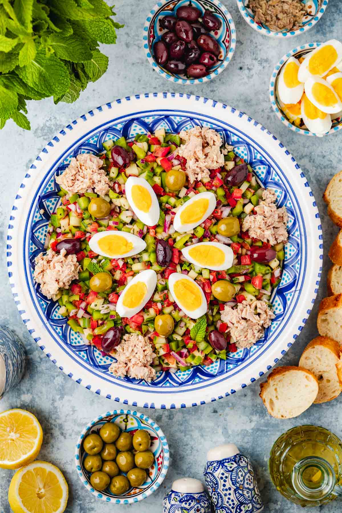 overhead photo of Slata Tounsiya Tunisian Salad in a serving bowl surrounded by lemon halves, mint, salt, pepper, olive oil, sliced bread, and bowls of green olives, kalamata olives, tuna and sliced hard boiled eggs.