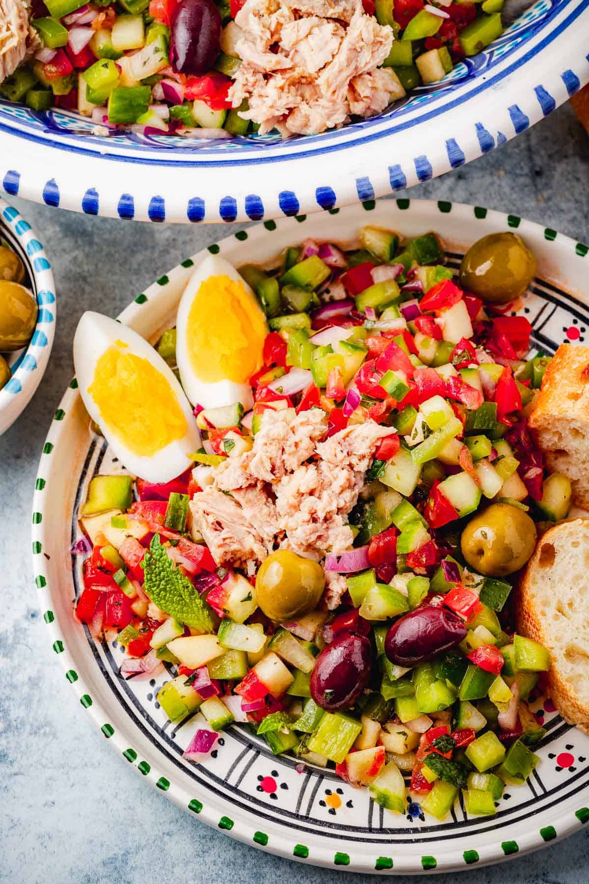 close up of a serving of Slata Tounsiya Tunisian Salad on a plate with slices of crusty bread in front of a serving bowl of the entire tunisian salad.