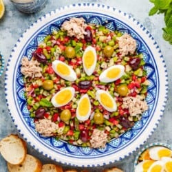 Slata Tounsiya Tunisian Salad in a serving bowl surrounded by lemon halves, mint, salt, pepper, olive oil, sliced bread, and bowls of green olives, kalamata olives, tuna and sliced hard boiled eggs.