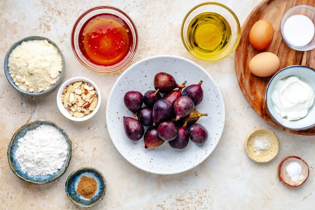 Ingredients for fig cake, including almonds, honey, olive oil, greek yogurt, eggs, salt, cinnamon, baking soda, flour, figs, and sugar.