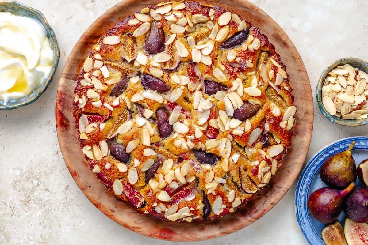 Overhead shot of fig cake on a wooden platter with fresh figs, sliced almonds, and Greek yogurt on the side.