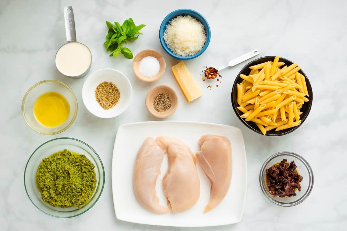 ingredients for pesto chicken pasta including chicken breasts, penne pasta, italian seasoning, salt, pepper, red pepper flakes, olive oil, heavy cream parmesan cheese, sun dried tomatoes, basil pesto, and basil leaves.