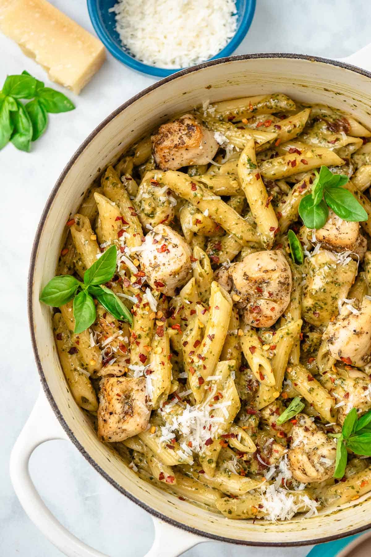 pesto chicken pasta garnished with basil leaves in a pot, next to a small bowl of grated parmesan, a small piece of parmesan, and basil leaves.