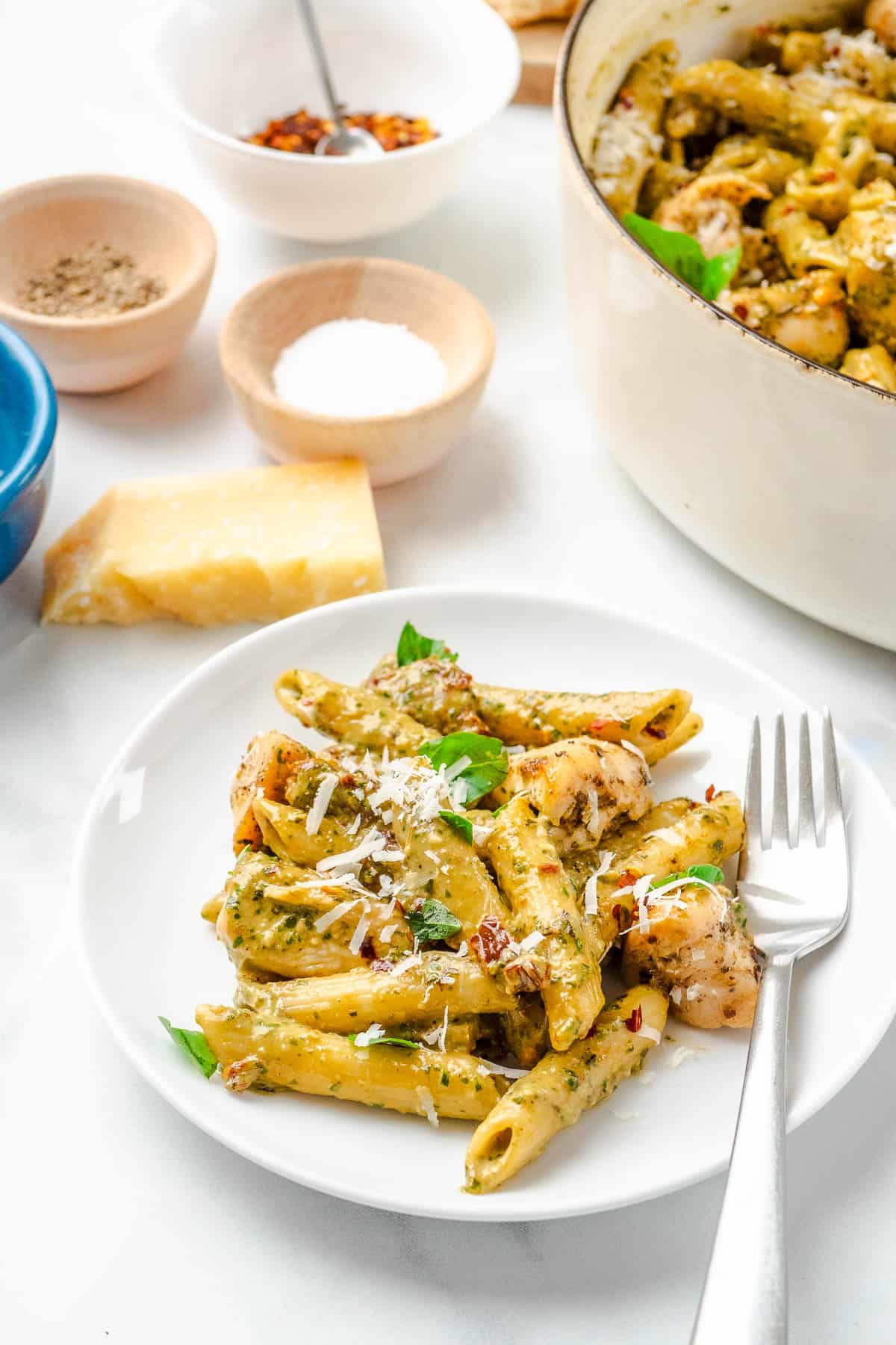 a serving of pesto chicken pasta on a white plate with a fork next to bowls salt, pepper, and red pepper flakes, a small piece of parmesan cheese, and a pot of pesto chicken pasta.