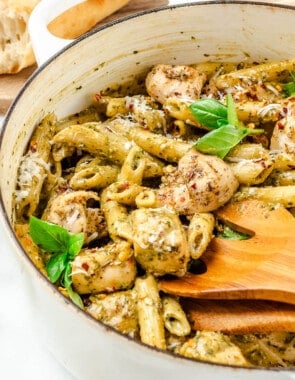 a pot of pesto chicken pasta with wooden serving utensils, next to bowl a bowl of red pepper flakes, basil leaves, and a cutting board with crusty bread.