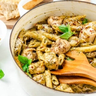 a pot of pesto chicken pasta with wooden serving utensils, next to bowl a bowl of red pepper flakes, basil leaves, and a cutting board with crusty bread.