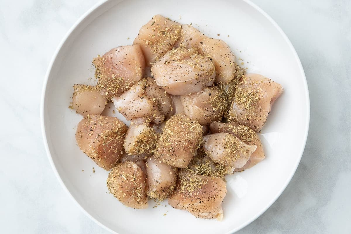Pieces of raw chicken breast on a white plate, sprinkled with italian seasoning.