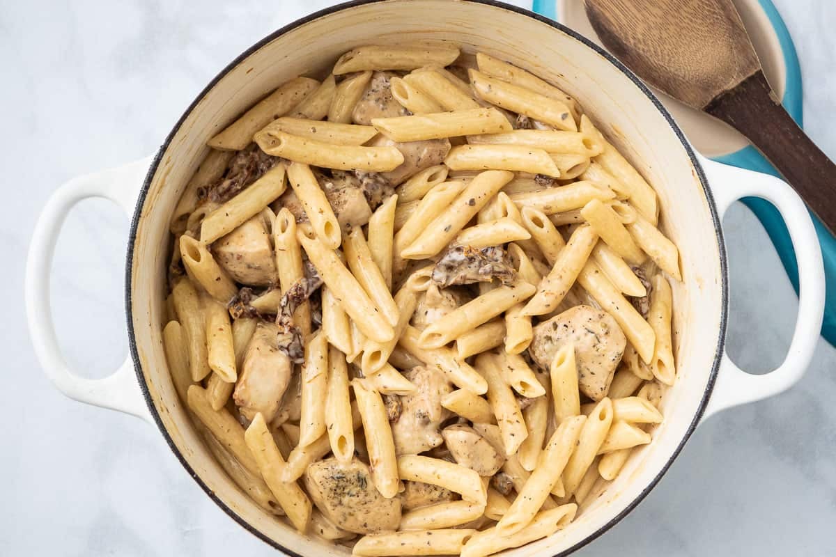 cooked penne pasta and pieces of chicken breast mixed with heavy cream and parmesan in a pot next to a wooden serving spoon on a spoon rest.