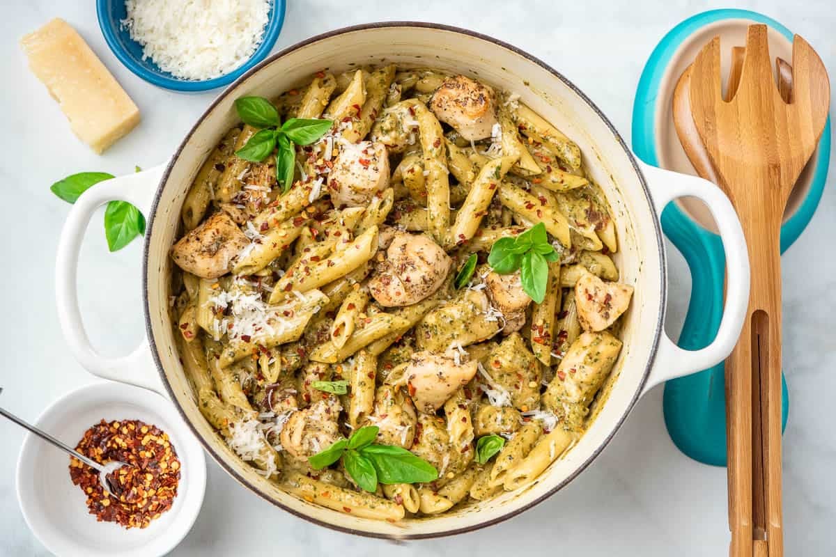 a pot of pesto chicken pasta next to wooden serving utensils, bowls of grated parmesan cheese and red pepper flakes, a piece of parmesan cheese and basil leaves.