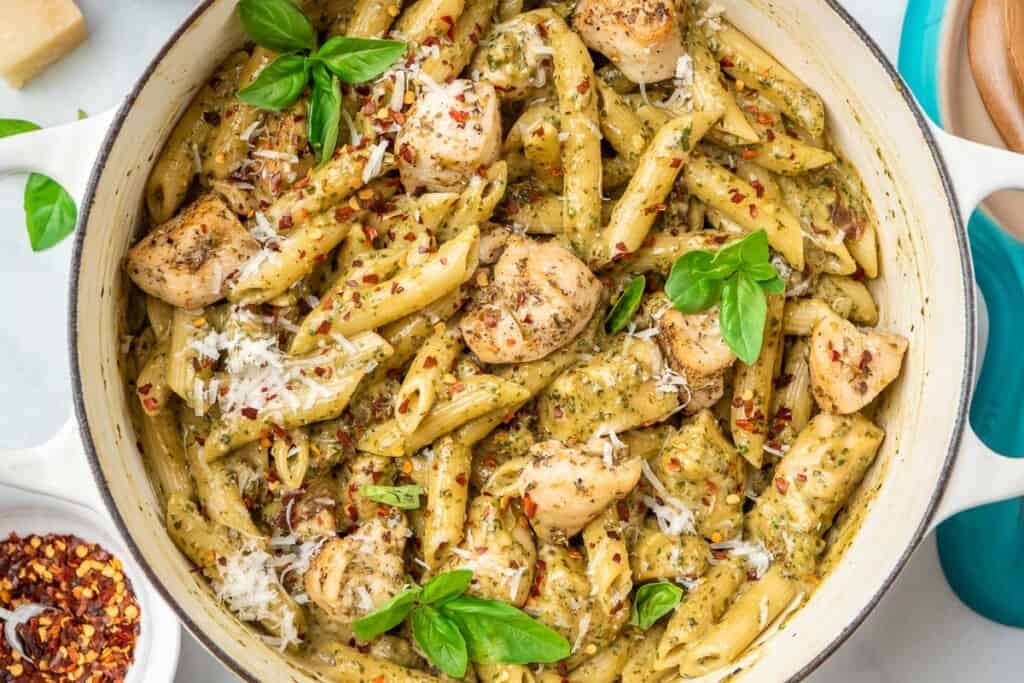 pesto chicken pasta garnished with basil leaves in a pot, next to a small bowl of red pepper flakes.