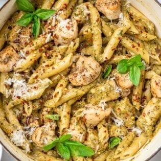 pesto chicken pasta garnished with basil leaves in a pot, next to a small bowl of red pepper flakes.