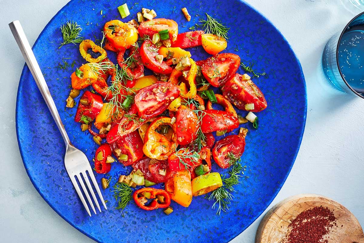 close up of a serving of bell pepper salad on a blue plate with a fork next to a bowl of ground sumac and a glass of water.