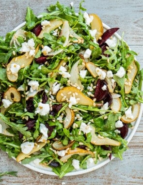 beet and goat cheese salad on a serving platter next to a jar of honey balsamic vinaigrette with a whisk and a bowl of pine nuts.