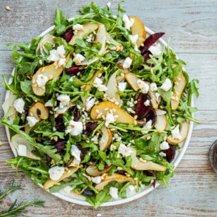 beet and goat cheese salad on a serving platter next to a jar of honey balsamic vinaigrette with a whisk and a bowl of pine nuts.