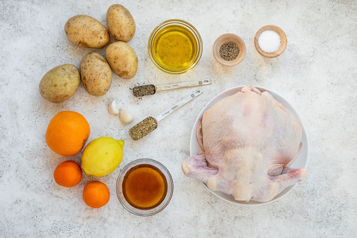 ingredients for roast spatchcock chicken including a whole chicken, potatoes, clementine oranges, naval orange, lemon, garlic, salt, pepper, olive oil, dried thyme and dried oregano.