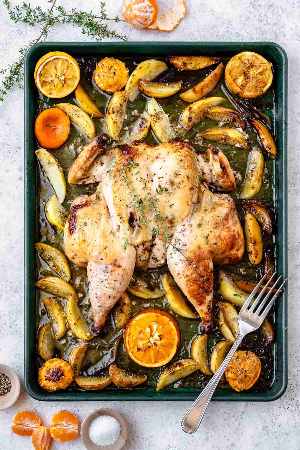 roast spatchcock chicken on a bed of potato wedges and citrus pieces on a sheet pan with a fork surrounded by bowls of salt and pepper, sprigs of thyme and peeled clementine oranges.