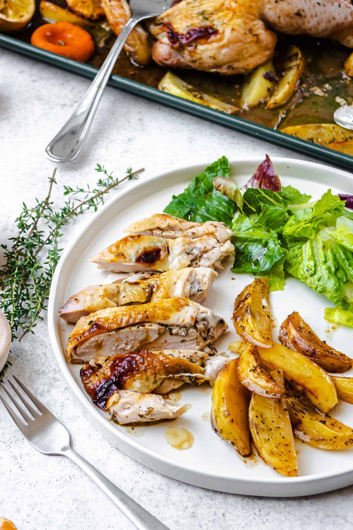 a piece of sliced roast spatchcock chicken with potato wedges and a salad on a plate next to a fork, sprigs of fresh thyme, and the sheet pan with the rest of the chicken and potato wedges.