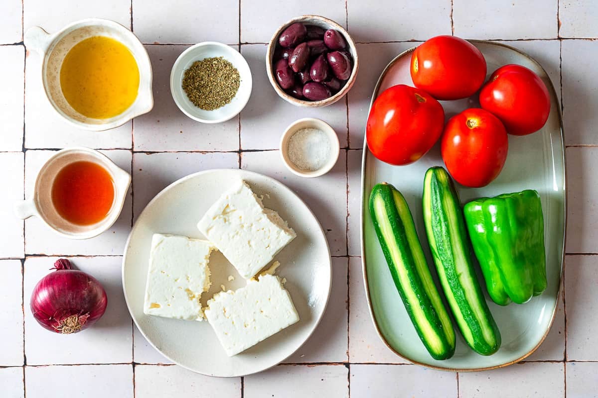 Ingredients for Greek salad including cucumbers, tomatoes, green pepper, feta cheese, red onion, kalamata olives, salt, oregano, olive oil, and red wine vinegar.