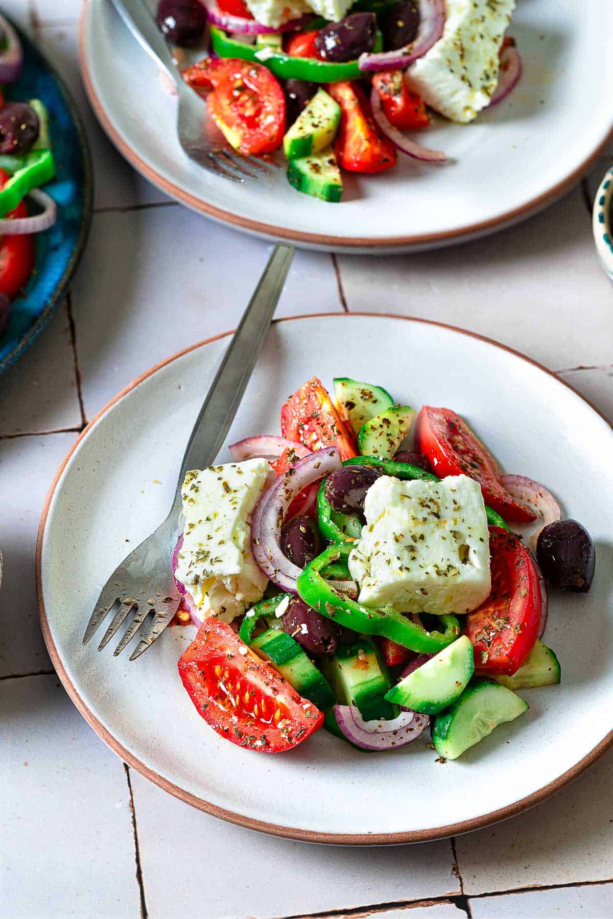 A serving of Greek salad one a white plate with a fork, with another in the background.
