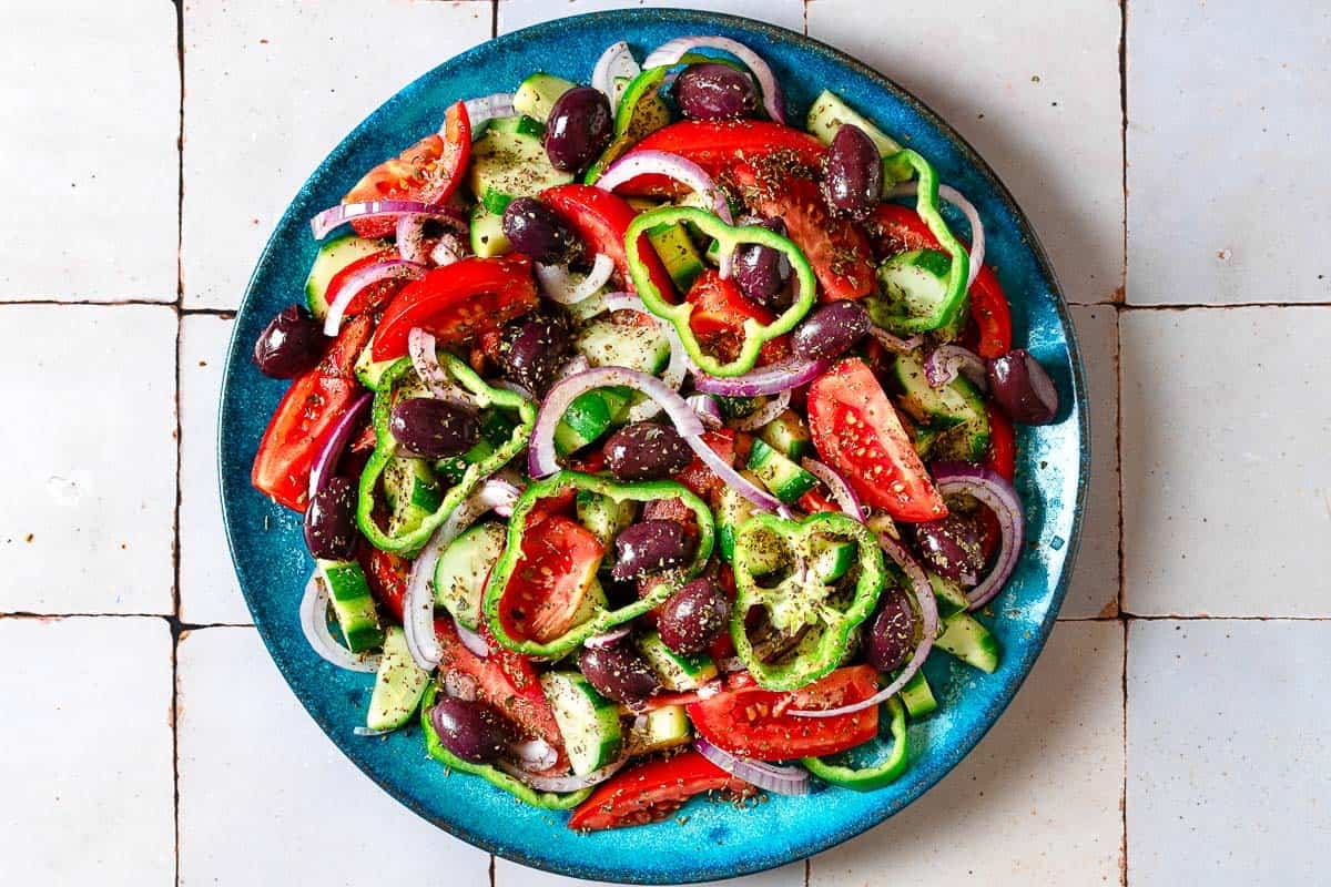 Overhead photo of a Greek salad on a blue serving platter before the blocks of feta cheese has been added.
